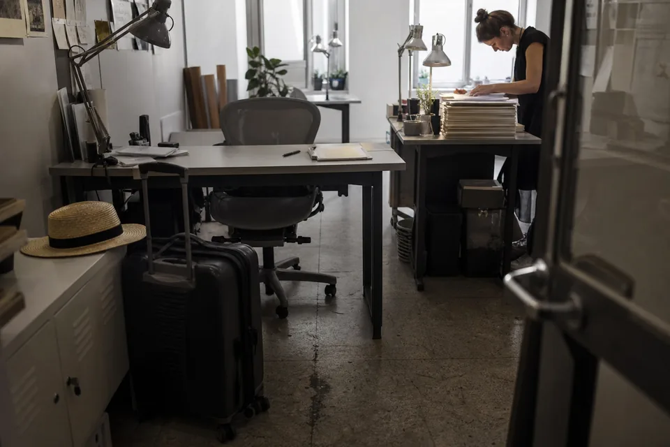 Empty desk with a suitcase beside it while a coworker at the next desk works through a stack of folders