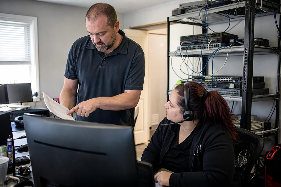 IT service manager holding a printed ticket list beside a technician wearing a headset in a small MSP office
